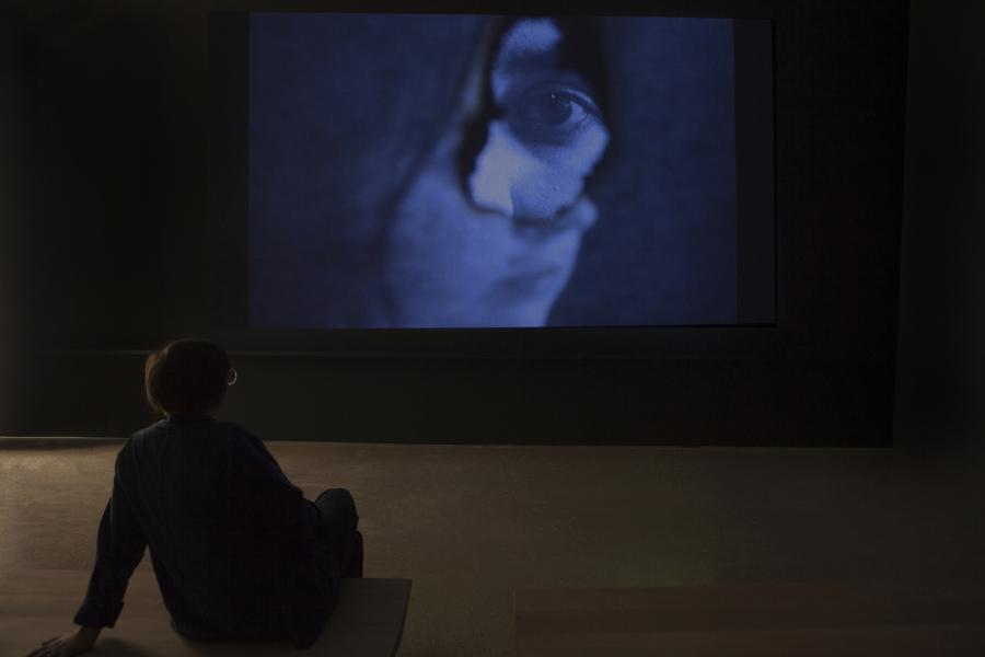 A visitor sits in a darkened space watching a video projection featuring a close-up of an eye through a fabric tear.