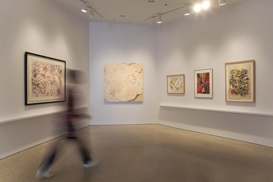 A gallery corner displaying abstract and expressive paintings and drawings on white walls, with a blurred figure walking past.