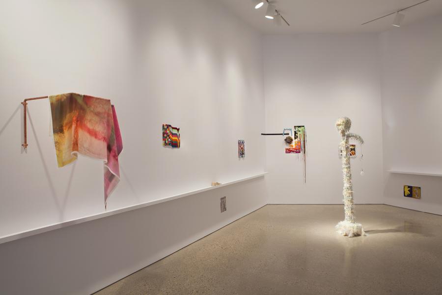 A wider gallery shot capturing various textile-based artworks along the walls, a central sculpture covered in feathery material, and a draped fabric piece on a copper rod.
