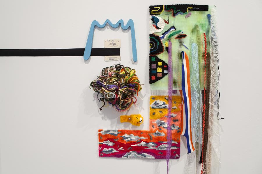 A mixed-media wall artwork featuring colorful cross-stitch elements, tangled multicolored threads, lace, ribbons, and a small plush lion. A blue wooden element resembling a crown is mounted above the composition, accompanied by vintage-looking price tags.