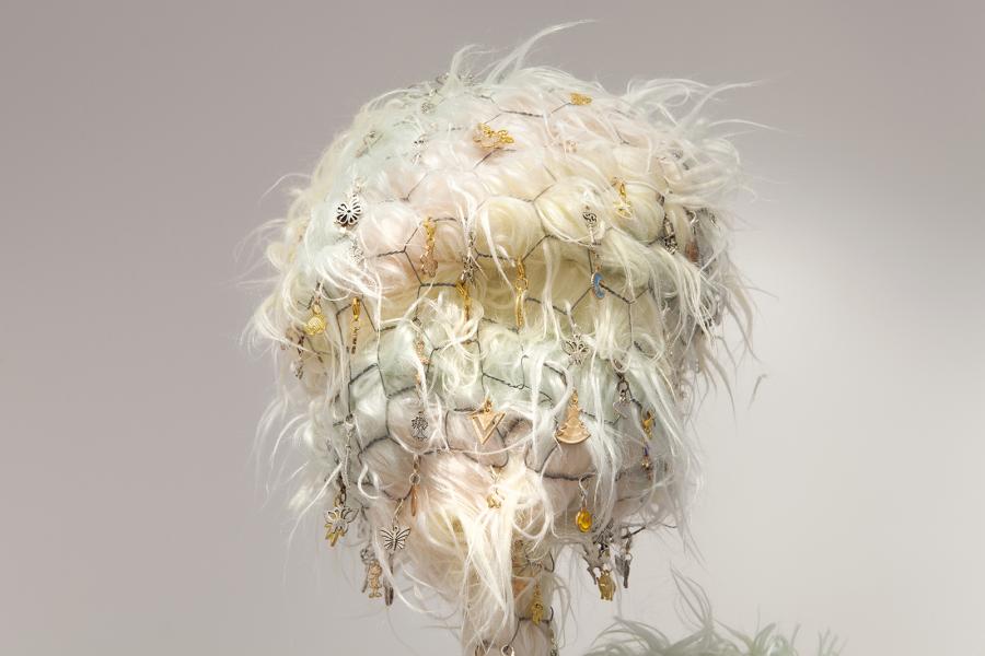 A close-up of the feathery, textured sculpture with various charms attached, including symbols like butterflies, hearts, and trees, intricately woven into the soft material.