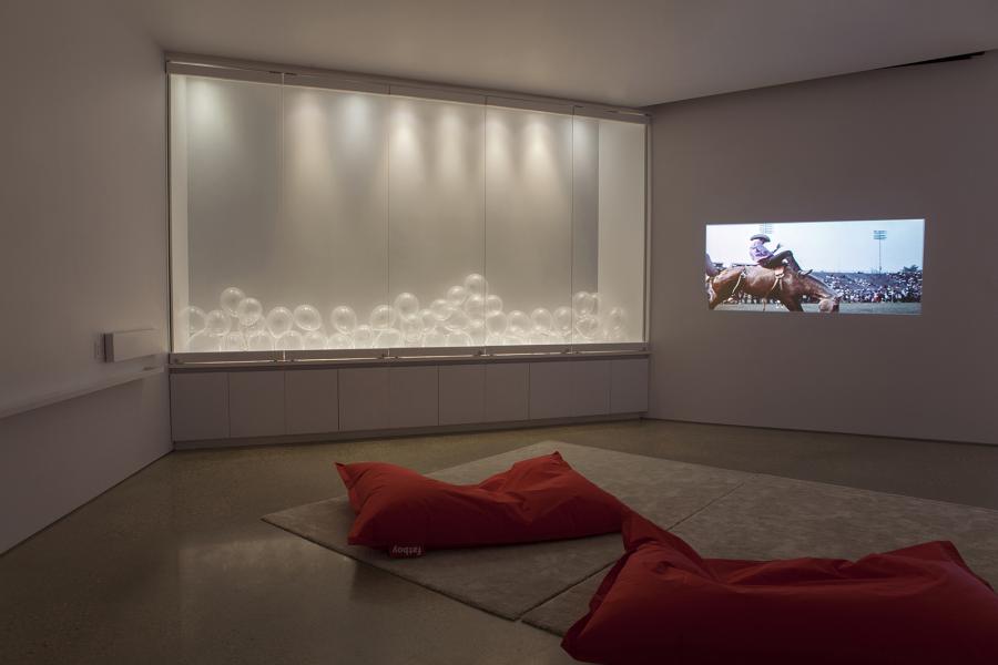 A gallery installation with a projection of a rodeo scene, a display of clear balloons in a lit cabinet, and red beanbag seating on a gray rug.