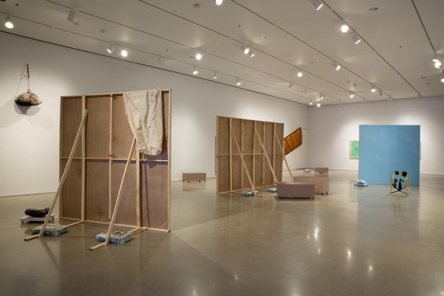A wide view of the gallery space, showing multiple freestanding wooden panels, scattered sculptural elements, and a vibrant blue partition, suggesting an installation in progress.