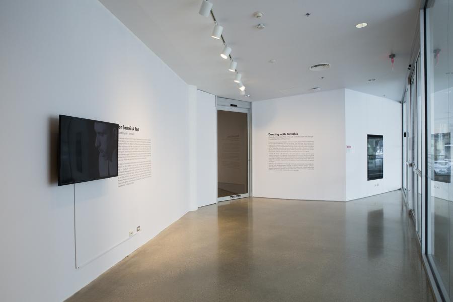 A gallery exhibition space with a video screen mounted on the wall, displaying Jon Sasaki’s A Rest, with exhibition texts including Dancing with Tantalus visible on the walls.