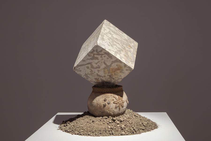 A sculptural piece consisting of a textured stone cube balanced atop a ceramic vessel, surrounded by soil on a white pedestal.