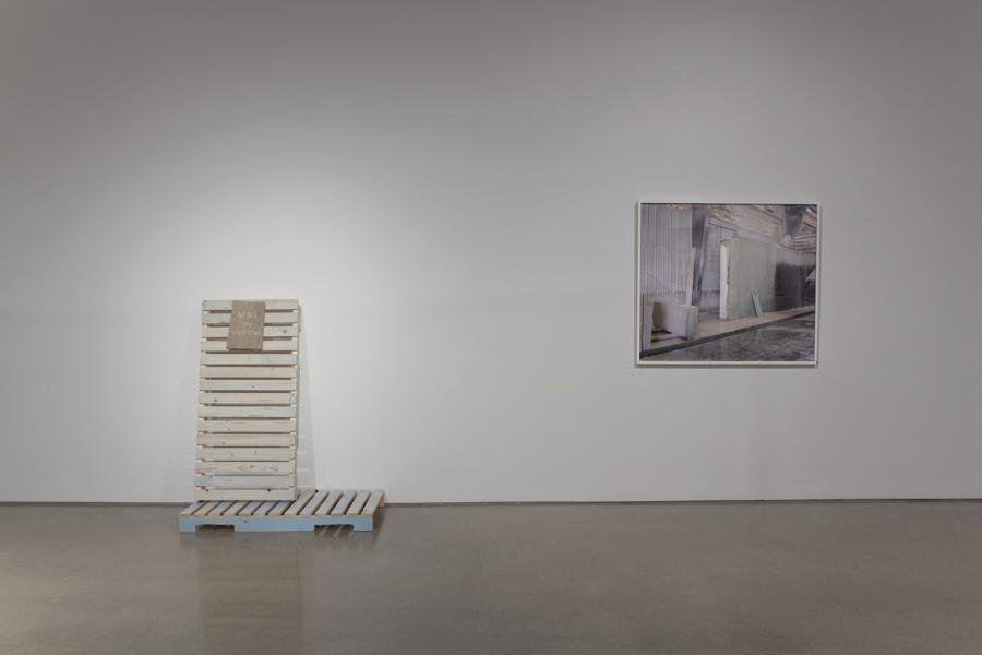 A wooden pallet installation labeled Notes on Harvesting, positioned beside a framed photograph of an industrial setting on a white gallery wall.