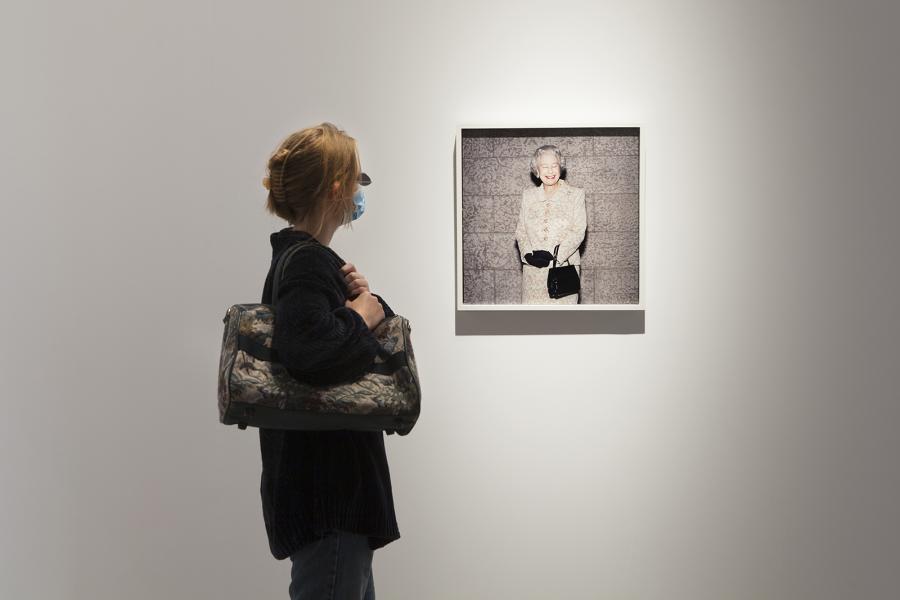 A visitor wearing a mask observes a framed photograph of an elegantly dressed elderly woman with a smile, displayed on a white gallery wall.