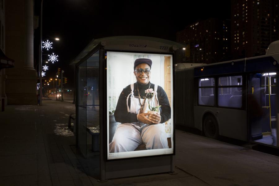 A night scene of a bus shelter with an illuminated portrait of a smiling individual in overalls, holding a potted plant, set against a backdrop of festive street lights.
