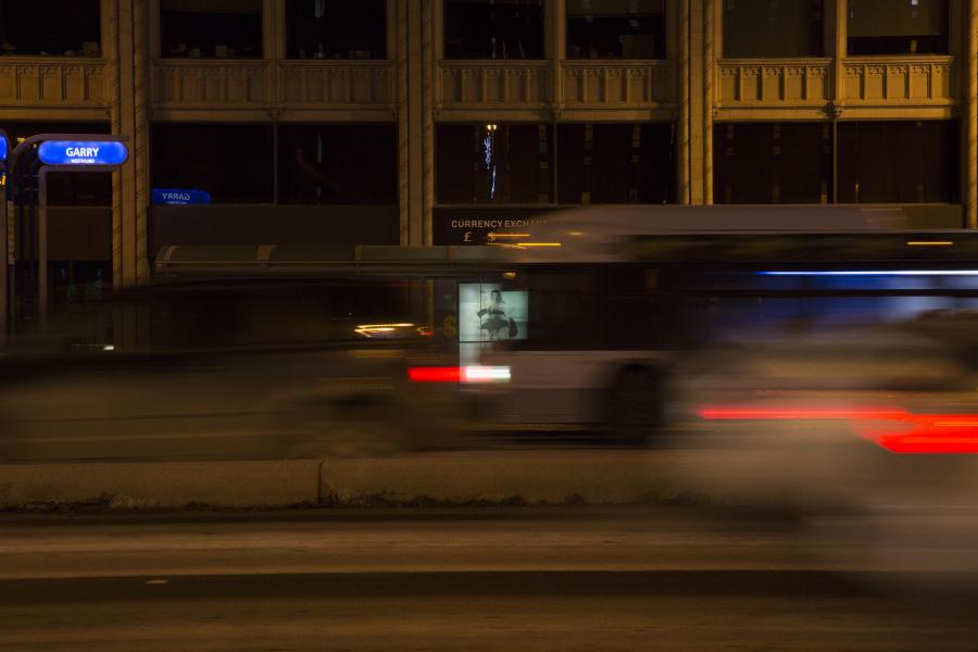 A city street at night with blurred motion from passing vehicles, partially revealing a lit bus shelter ad.