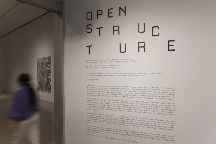 Exhibition title wall for Open Structure with curatorial text, partially obscured by a blurred figure walking past.