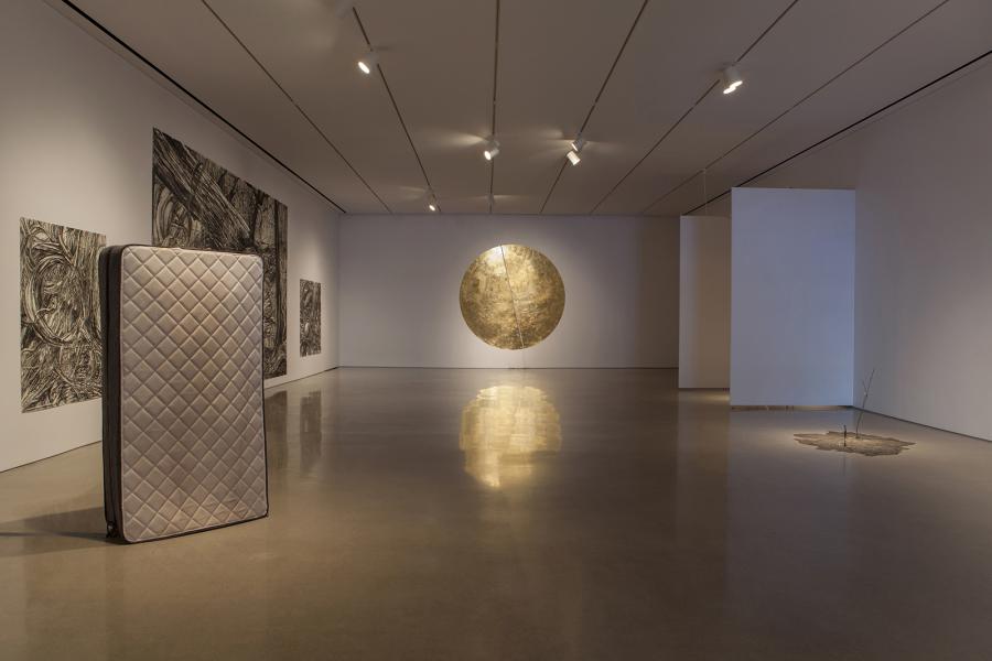 Wide gallery view showcasing a mattress, large charcoal drawings, a glowing circular artwork, and a fragmented floor installation.
