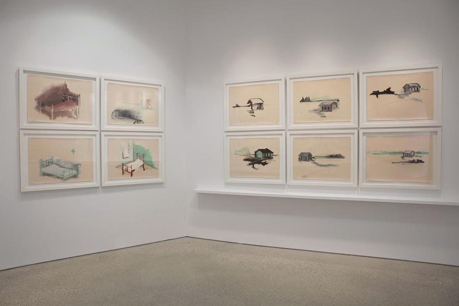 A gallery wall displaying a grid of framed drawings depicting isolated beds and buildings, rendered in soft washes of color with gestural ink lines.