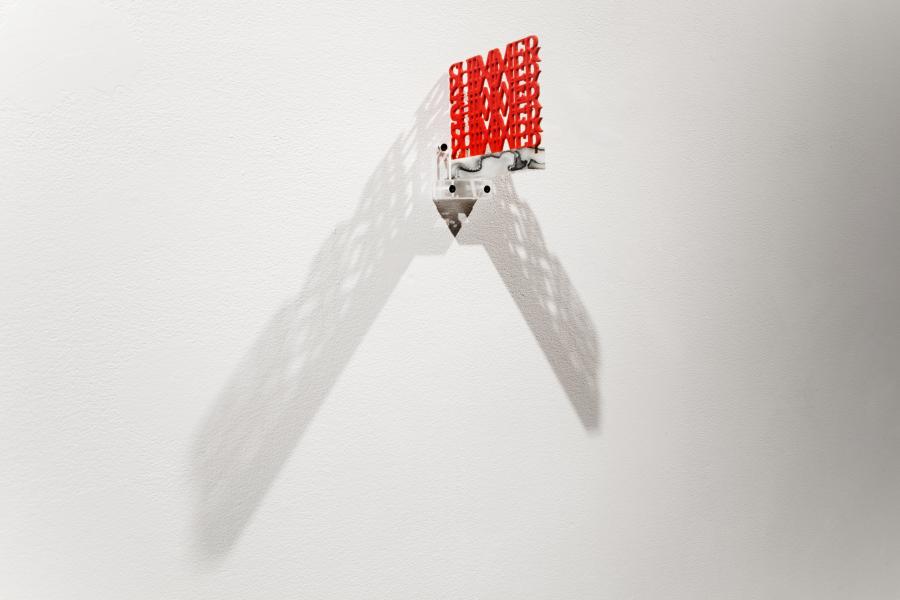 A small red plastic text sculpture is mounted on the gallery wall, projecting shadows that extend outward in a V-shape. The text is densely layered, creating depth.