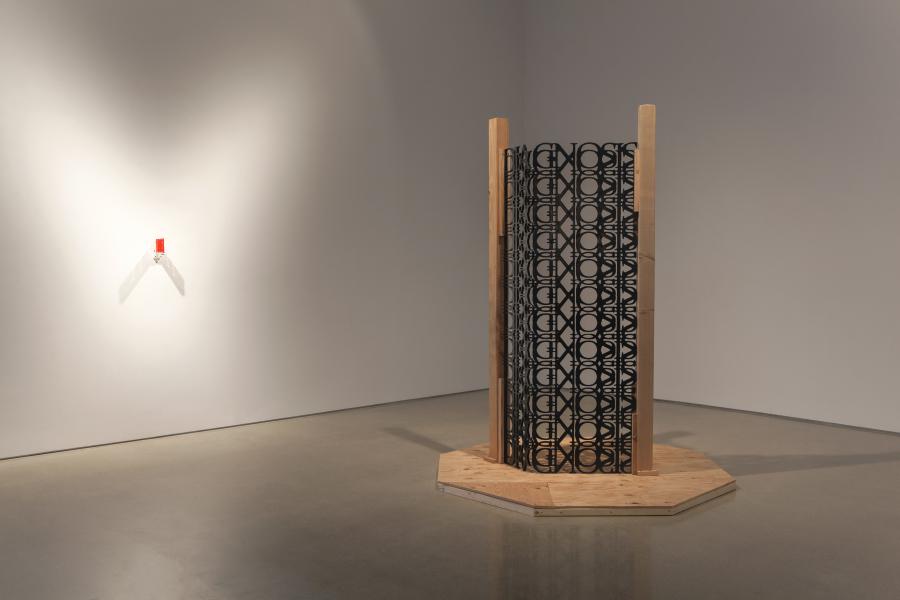 A black metal gate with intricate geometric cut-out patterns stands on a wooden platform. The gate casts a complex shadow on the floor, emphasizing its detailed design.