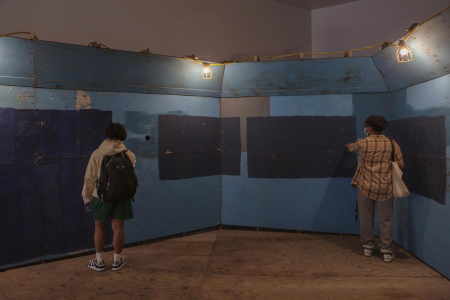 Two visitors observing and interacting with a blue wooden installation with painted rectangular patterns.