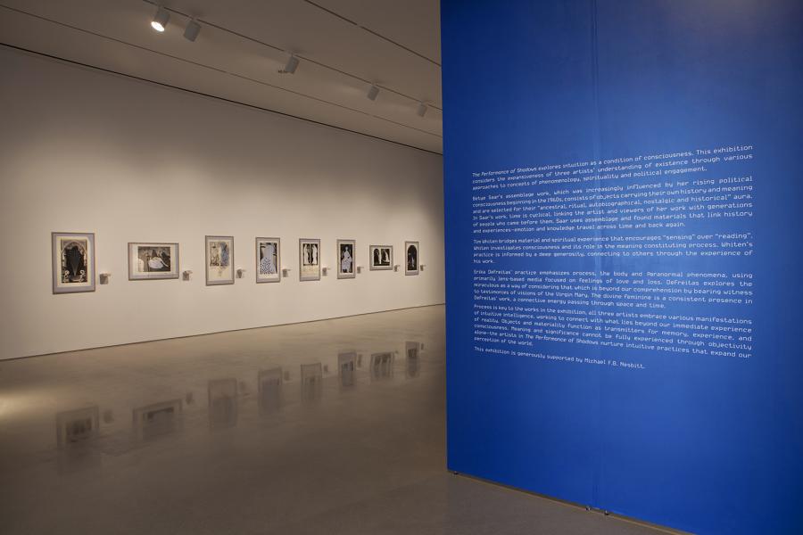 A gallery wall displaying framed black-and-white artworks, with a large blue text panel providing curatorial context for the exhibition.