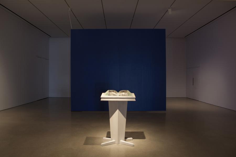 A frontal view of a book sculpture on a wooden pedestal, set against a striking blue exhibition wall.