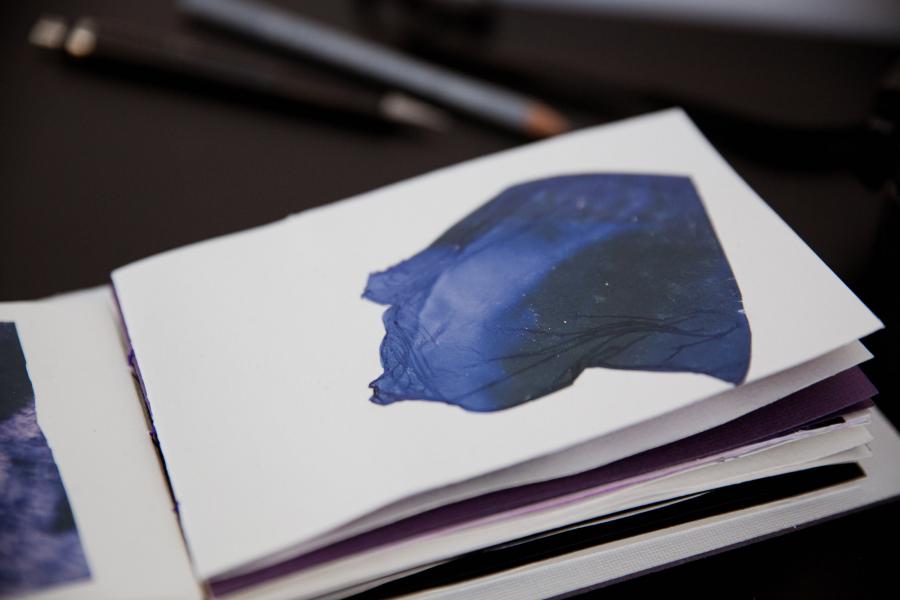 [10:14 AM] Cailyn Harrison An open artist book showing a deep blue abstract image on a white page, with drawing tools blurred in the background.
