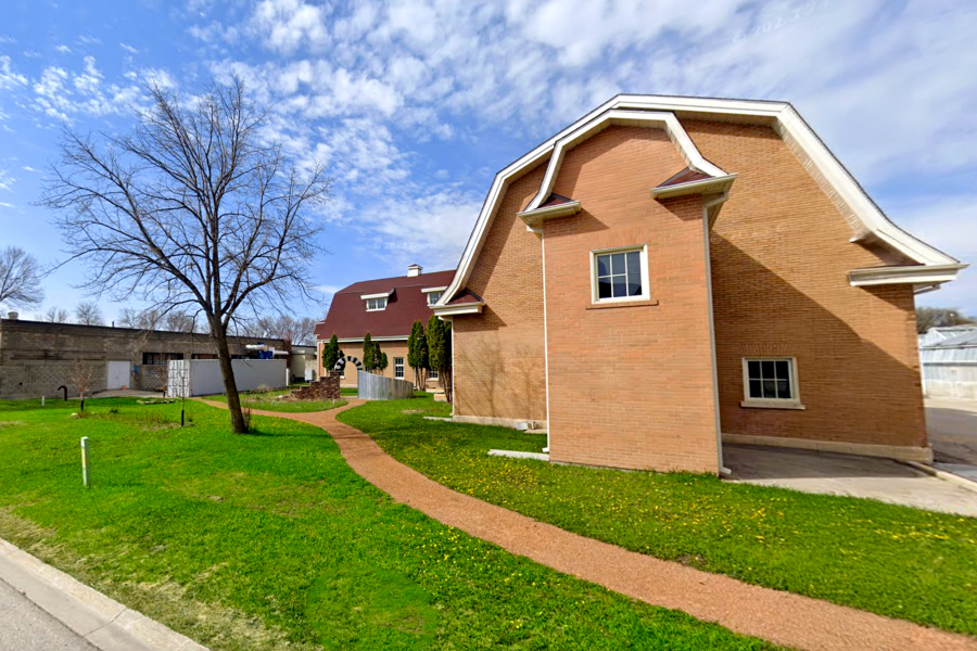 Exterior view of the Art Barn at the University of Manitoba, showing a red-roofed brick building with a winding path and green lawn under a partly cloudy sky.