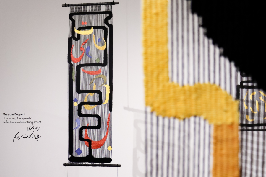 Detail of a hanging textile work by Maryam Bagheri titled Unwinding Complexity: Reflections on Disentanglement. The work features densely embroidered calligraphic forms in red, blue, yellow, and green on black and white vertical threads. Wall text includes the title in English and Farsi.