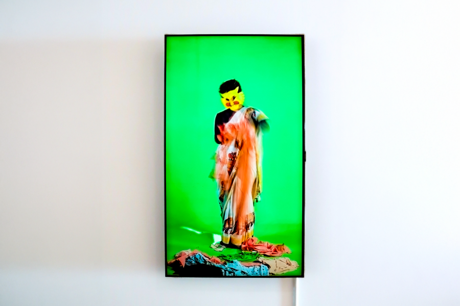 Video still from Laila Fazal's performance-based work featuring a figure wearing a stylized cat mask, standing in front of a vivid green background while draped in layered fabric.