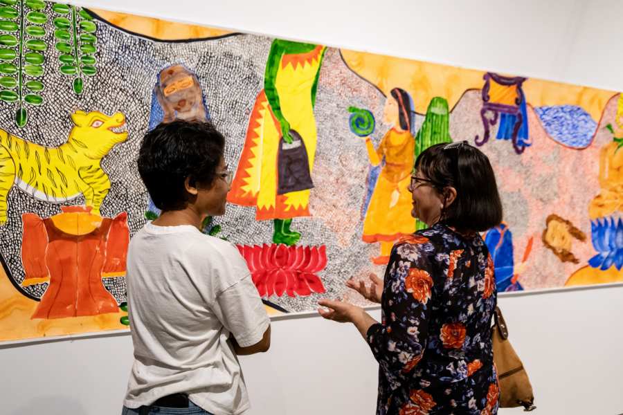 Two viewers engaging with Laila Fazal’s mural “Woven Roots,” discussing the vivid scenes of tigers, figures, and traditional garments depicted in the colorful panoramic artwork.