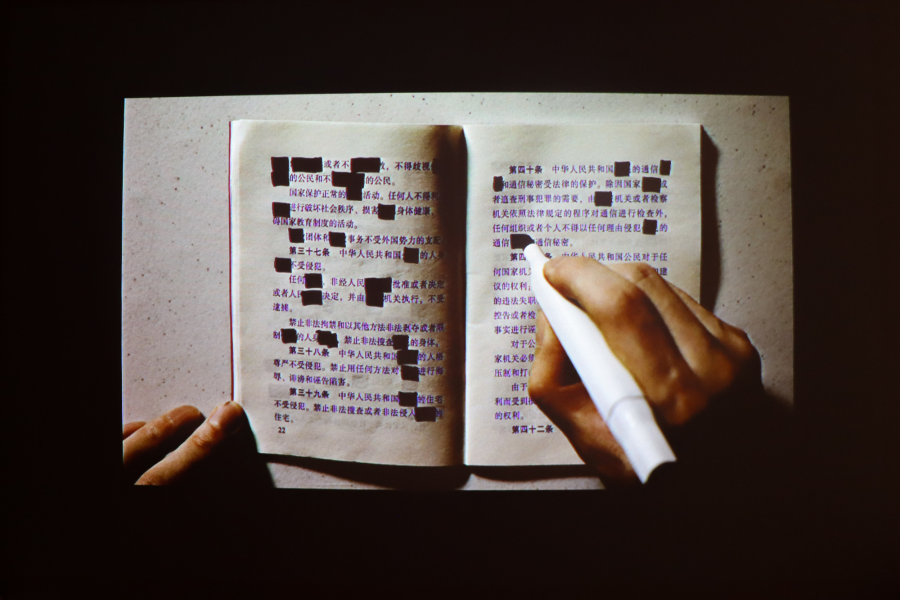 A projection of a book spread covered in Chinese characters, with some words redacted in black marker. A hand with a white pen is in the process of crossing out more text.