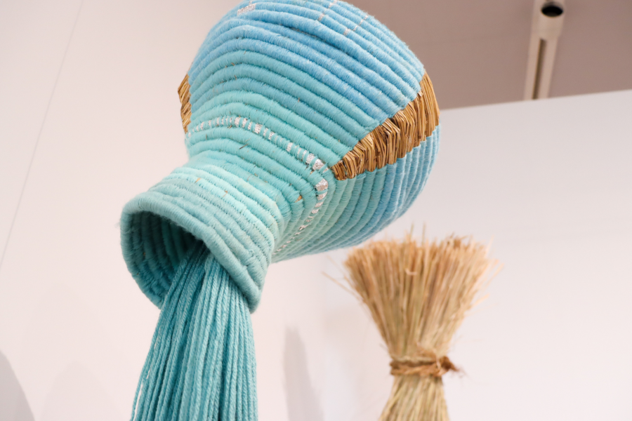 Detail view of a hanging vessel wrapped in soft turquoise yarn with embedded straw textures, combining textile processes and sculptural forms in Shawl’s installation.