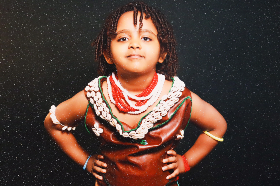 Studio portrait of Shawl's child in a shell- and bead-embellished garment, hands on hips, set against a dark glittery background—part of Shawl’s exploration of cultural memory and identity.