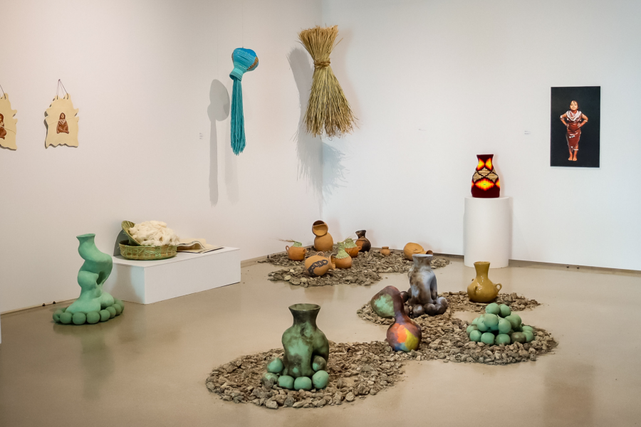 Wide installation view of Shawl’s MFA exhibition featuring clay vessels, textile works, and photo-based ceramics arranged on platforms and earth-like surfaces in a gallery setting.