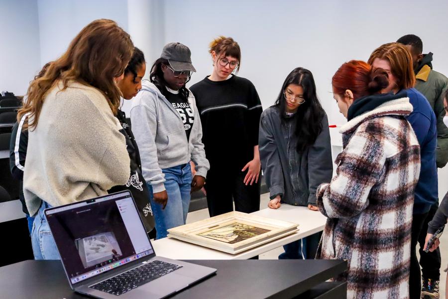  group of students gathers around a table to closely examine framed artworks in a classroom setting, with a laptop displaying a digital image in the foreground.