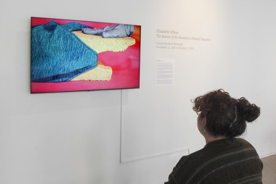 A viewer seated in front of Elizabeth Milton’s video installation at the School of Art Gallery, University of Manitoba. The screen shows a close-up of sponge-like material supporting a turquoise fabric and a gloved hand against a red background. On the adjacent wall, exhibition text reads: Elizabeth Milton: The Bottom of the Fountain is Painted Turquoise, curated by Blair Fornwald, November 21, 2024 to February 1, 2025.