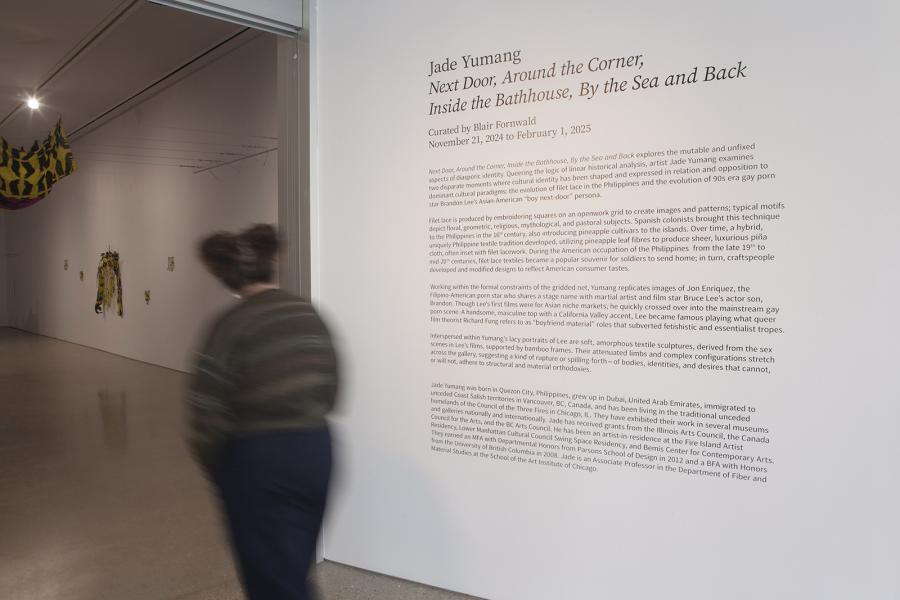 A visitor walks past the exhibition title wall text, which introduces Jade Yumang’s show Next Door, Around the Corner, Inside the Bathhouse, By the Sea and Back. Inside the gallery, yellow-and-pink fabric sculptures hang from the ceiling and walls.