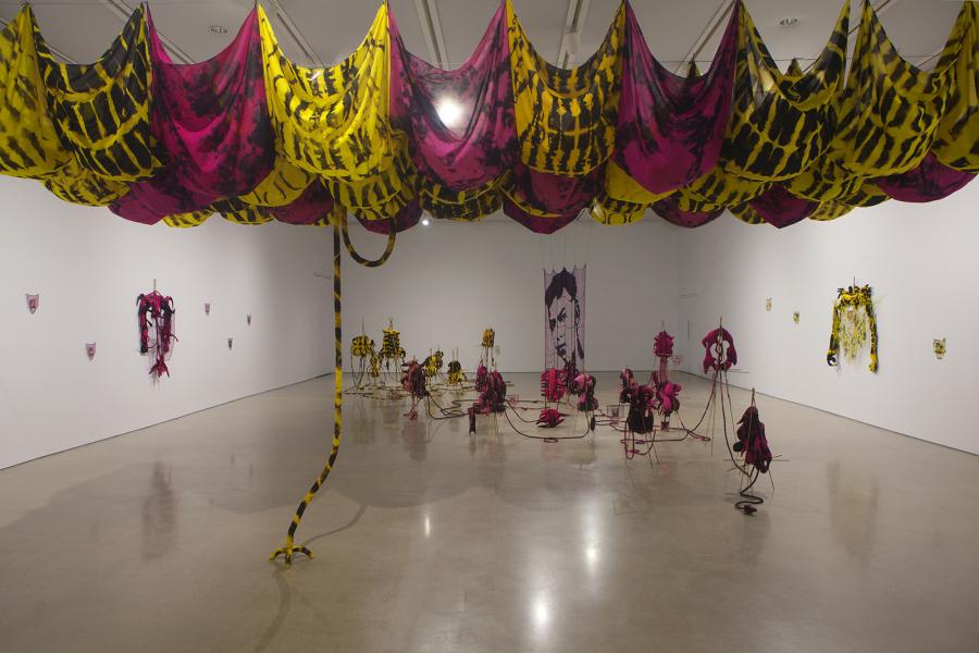 Wide view of the gallery installation showing a large canopy of yellow-and-pink tie-dyed fabric suspended from the ceiling, with clusters of red and yellow textile sculptures connected by cords across the floor. A woven portrait of a face hangs at the far wall.