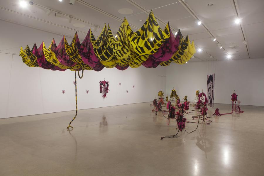Alternate angle of the installation featuring the yellow-and-pink fabric canopy overhead, textile sculptures arranged on the floor, and a portrait woven into netting displayed on the back wall.