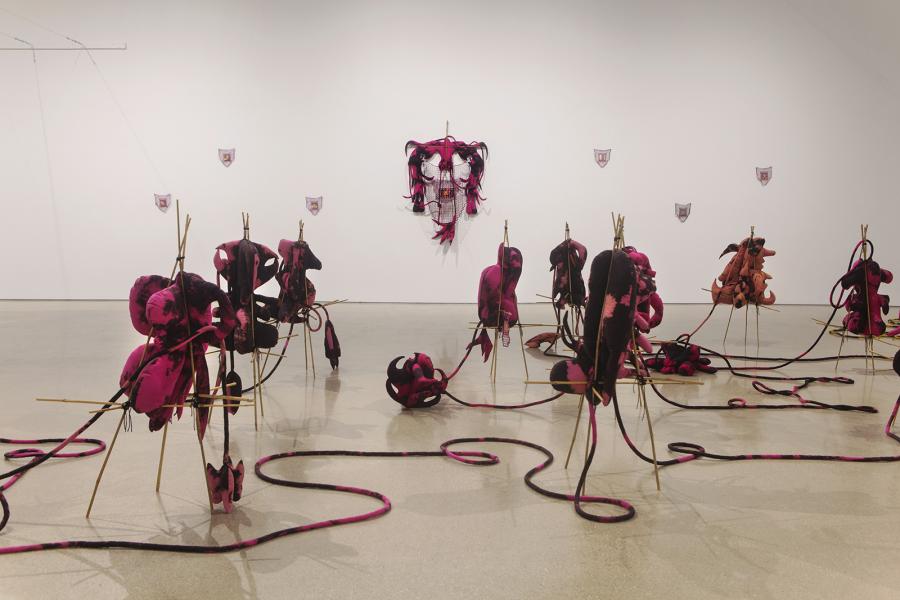 Installation of pink textile sculptures arranged in rows on the gallery floor, each tied to wooden supports, with cords trailing outward. On the wall hangs a mask-like woven textile piece surrounded by small shield-shaped works.