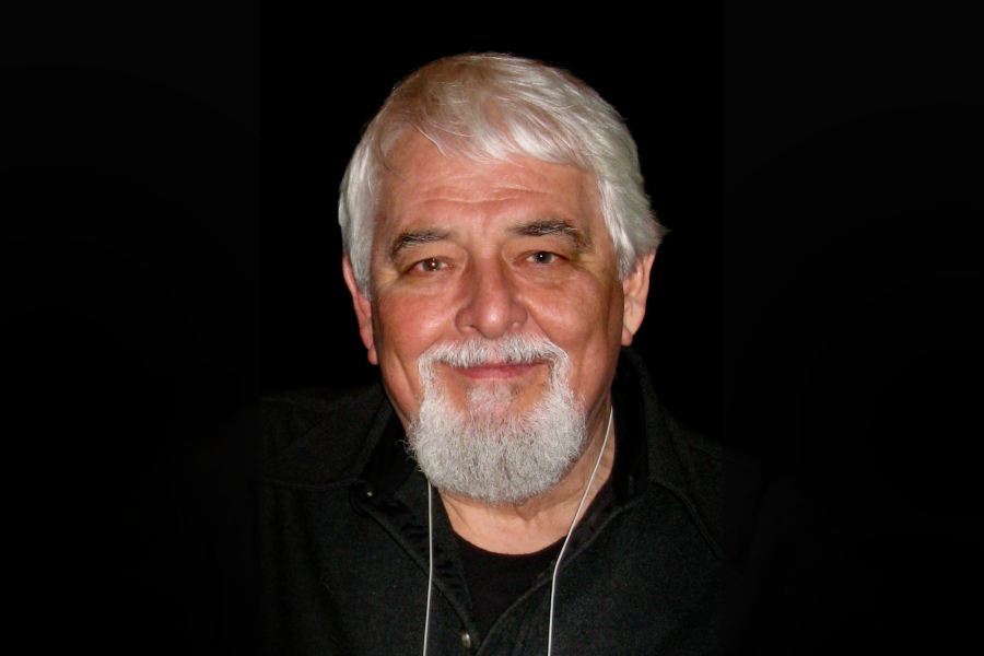 Portrait of Robert Archembeau with white hair and a full white beard, smiling warmly. He is wearing a black shirt against a solid black background.