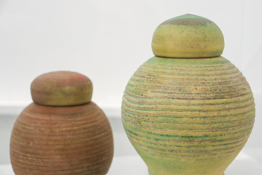 Two lidded ceramic vessels by Robert Archambeau displayed on a white surface. The foreground vessel is larger, with a softly ribbed texture and muted green glaze with earthy tones showing through. The smaller vessel behind it has a similar form with horizontal ridges and a warm reddish-brown surface. Both have rounded lids that echo the curves of the vessels, showcasing Archambeau’s subtle use of colour, texture, and form.