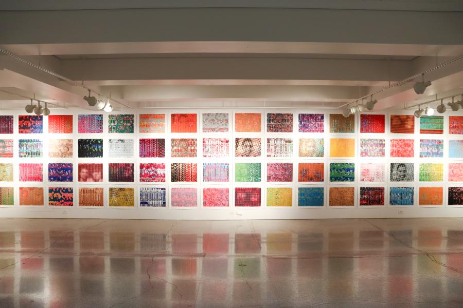 Wide view of a gallery wall covered with dozens of small, colorful square artworks arranged in a grid.