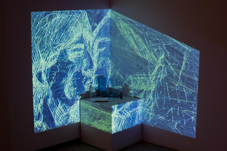 Projected blue and green abstract lines cover two gallery walls and a small pedestal installation.