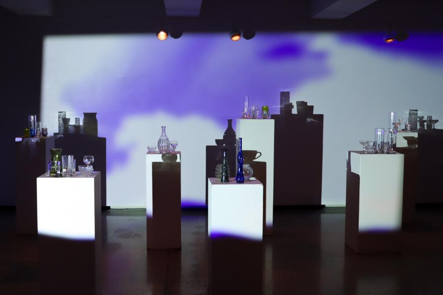 Installation view of ASTRAL: glass vessels and goblets arranged on white pedestals, casting tall shadows against a wall washed with purple-blue projected light.