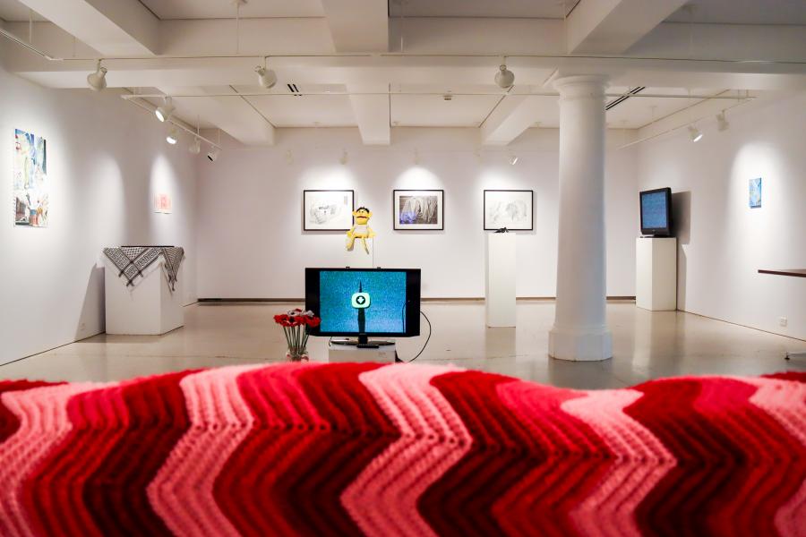Wide view of a gallery installation with video monitors, drawings on the walls, and a red crocheted textile in the foreground.