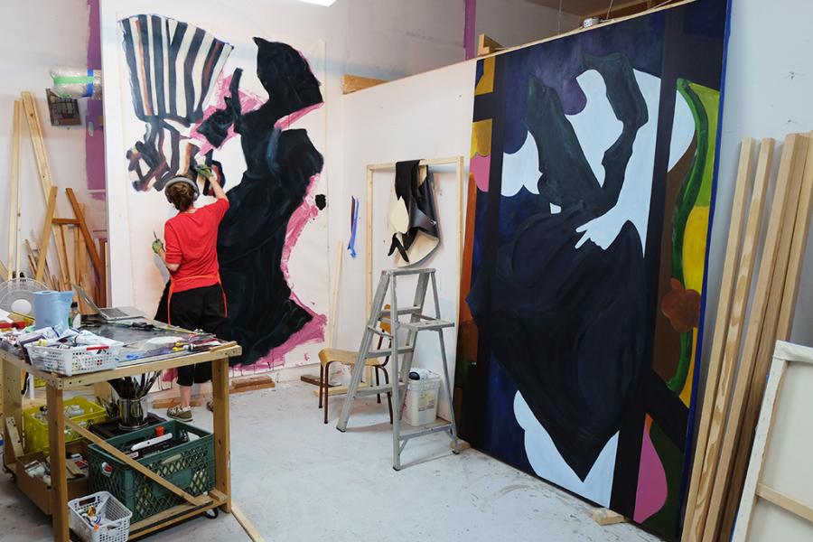 M.E. Sparks working in a studio beside a worktable, painting a large abstract canvas while another oversized painted panel leans against the wall.
