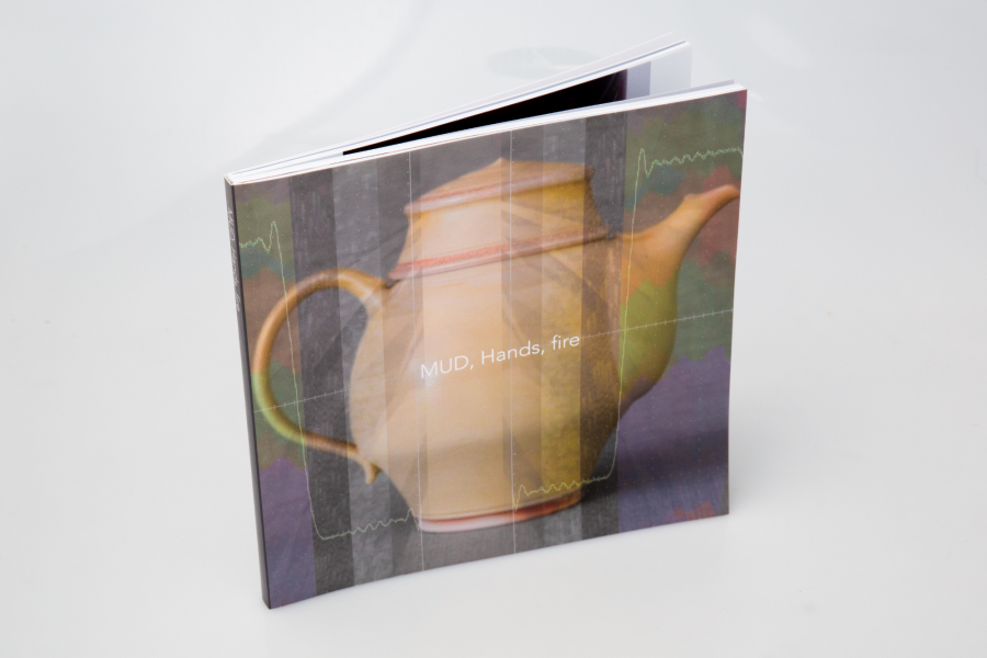 Small upright book on a white backdrop with the cover text “MUD, Hands, fire” over a layered image of a mug and waveform-like graphics.