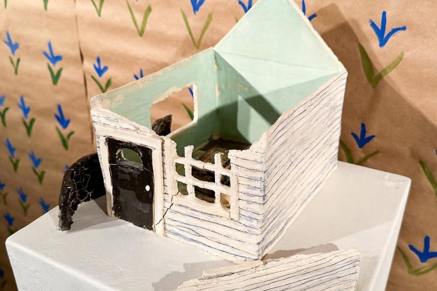 Small sculptural model of a house with pale blue interior walls, a black door, and broken exterior sections, displayed on a white pedestal against a patterned backdrop.
