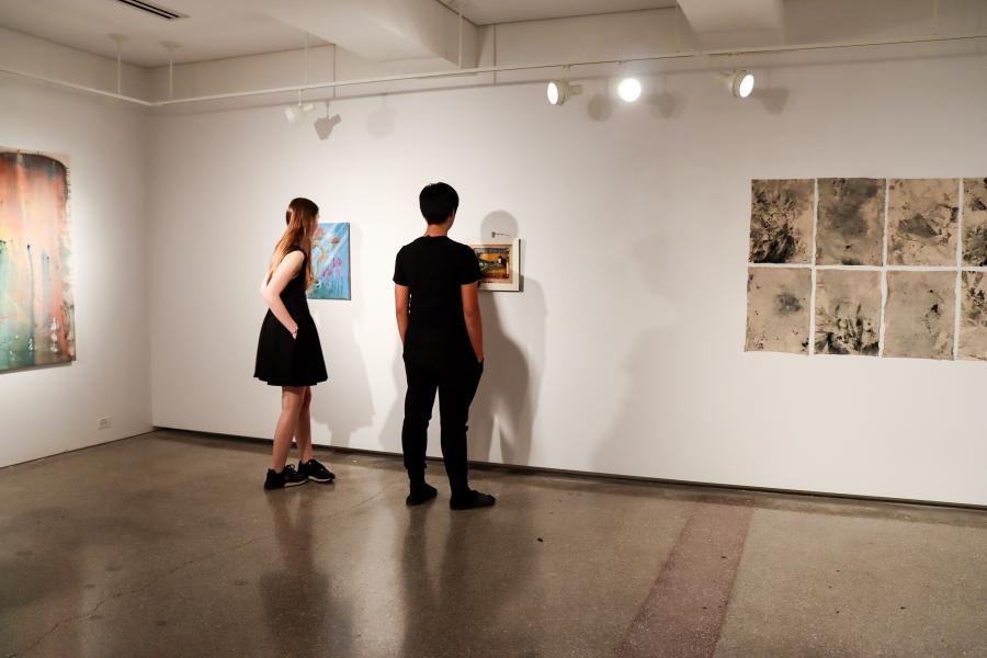 Two visitors stand in a gallery viewing small framed artworks on a white wall.
