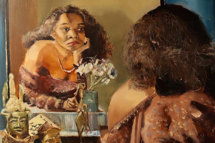 Painted figure reflected in a mirror beside small sculptural heads and flowers on a tabletop.