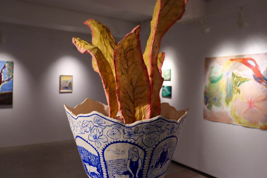 Large ceramic planter with blue illustrated scenes holds tall, leaf-like sculptural forms in a gallery space.