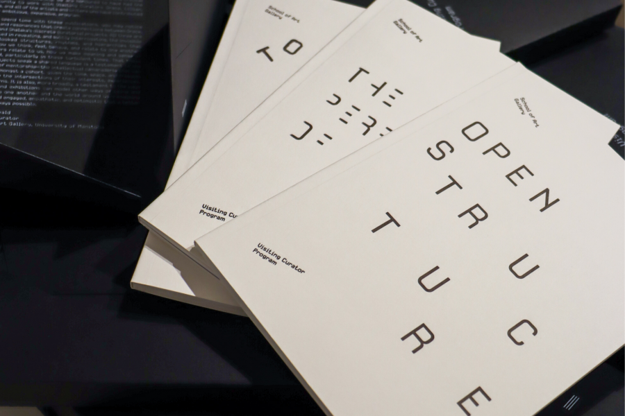 Angled close-up of stacked white booklets titled “Open Structure” (Visiting Curator Program) on a dark surface.