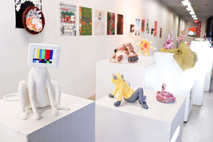 A hallway exhibition featuring a row of small sculptural works displayed on white pedestals, with colorful mixed-media pieces receding into the distance along a gallery corridor.